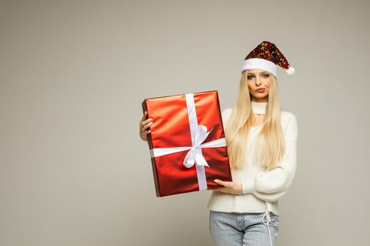 Stock photo of attractive blonde girl in Santa hat, white sweater and light blue jeans holding wrapped Christmas gift and looking at camera with doubtful facial expression.