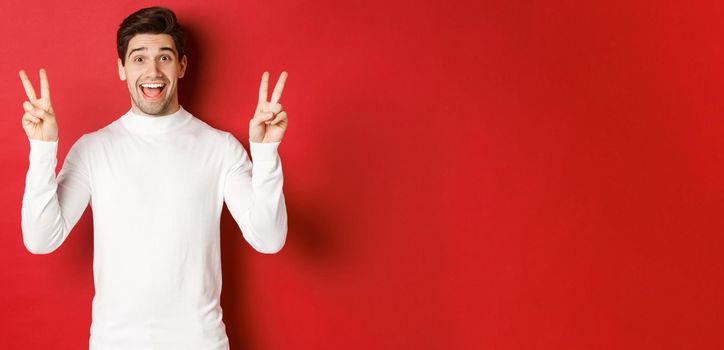 Concept of winter holidays, christmas and lifestyle. Handsome funny guy in white sweater, showing peace signs and smiling happy, standing against red background.