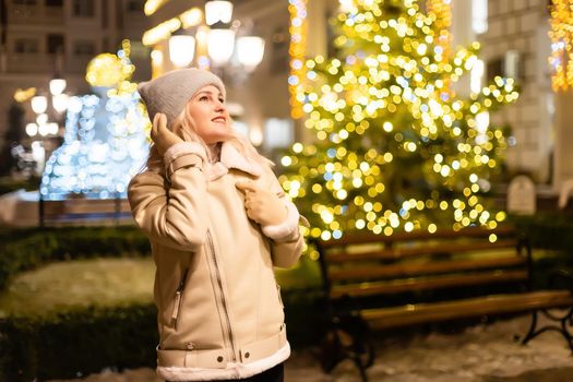 Outdoor photo of young beautiful happy smiling girl holding sparklers, posing in street. Festive Christmas fair on background. Model wearing stylish winter coat, knitted beanie hat, scarf.