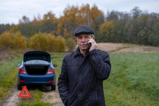 Adult pensioner calls technical assistance on smartphone after his faulty car outside city. Selective focus.