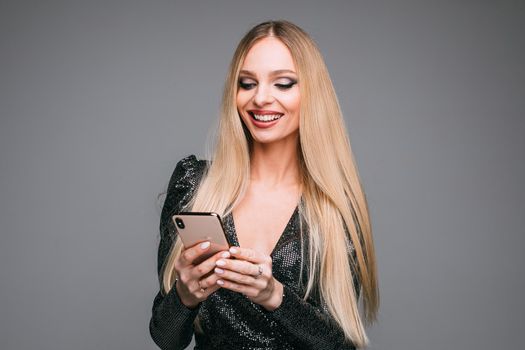 Stock photo of attractive blonde girl in sparkling cocktail dress using mobile to chat with friends and smiling. Isolate on grey.