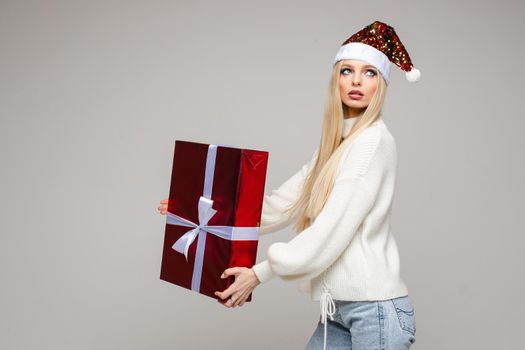 Pretty young lady standing in studio and holding large gift box, isolated on grey background. New Year holiday concept