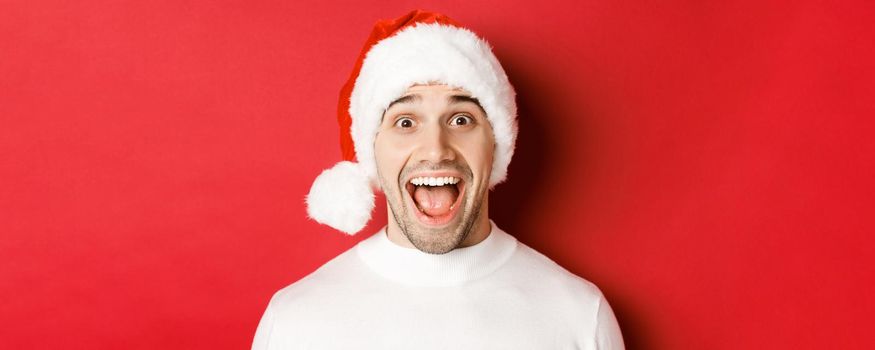 Concept of winter holidays, christmas and celebration. Close-up of surprised handsome man in santa hat, hear amazing new year promo offer, standing against red background.