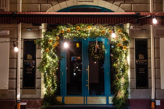 Stylish christmas tree with red berries, wreath on doors and wooden sleigh at front of store at holiday market in city street. Christmas street decor. Space for text. Rustic decoration.