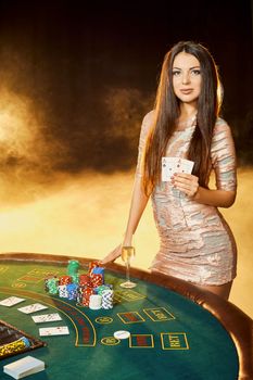 Gorgeous young woman in evening dress with two cards in hands standing near poker table with glass of champagne