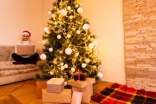 Pretty Young Woman in Trendy Winter Fashion Sitting on the Floor with Crossed Legs Holding Laptop Gadget in Front Christmas Tree Decoration, While Looking at the Camera.
