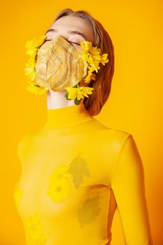 Slim female model in protective mask with fresh yellow flowers standing in transparent outfit on yellow background in studio