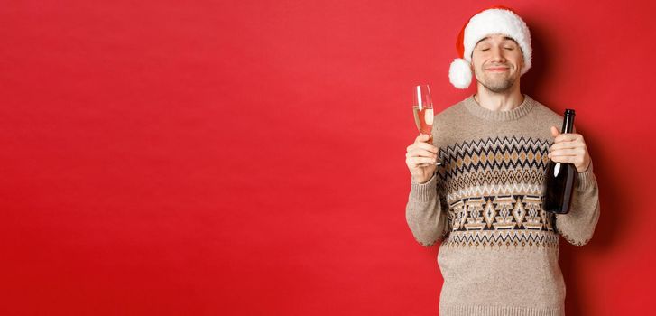 Concept of winter holidays, christmas and celebration. Image of pleased smiling man in santa hat and sweater, drinking on new year, holding bottle of champagne and filled glass, red background.