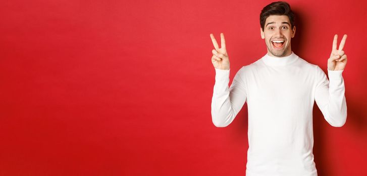 Concept of winter holidays, christmas and lifestyle. Handsome funny guy in white sweater, showing peace signs and smiling happy, standing against red background.