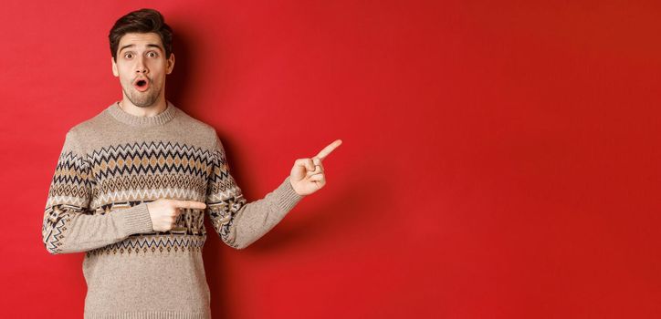 Concept of christmas celebration, winter holidays and lifestyle. Amazed handsome guy in xmas sweater pointing fingers right at logo, showing advertisement, standing over red background.