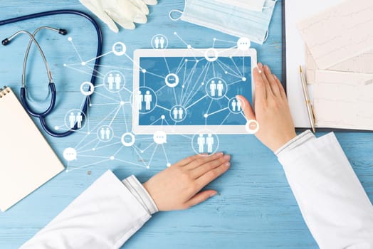 Top view of doctor hands with tablet computer. Therapist in white coat sitting at blue wooden desk. Examination and consultation in clinic. Medical application and online healthcare services.