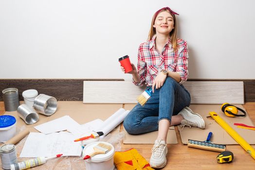 Dreamy girl painter relaxing on floor with cup of coffee. House remodeling and interior renovation concept with copy space. Young woman in red checkered shirt and jeans planning to redesign her home.