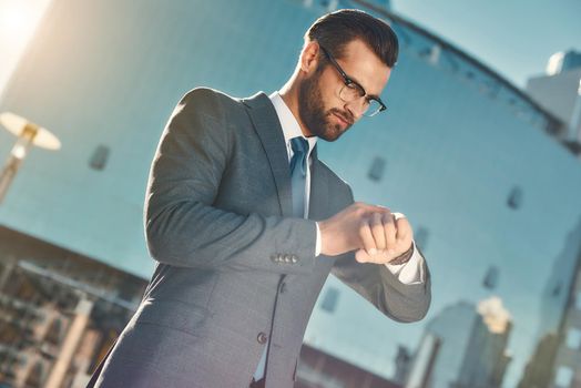 In a hurry. Young and handsome bearded businessman in eyeglasses and full suit checking the time while standing outdoors. Business concept. Time concept