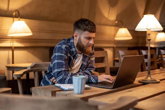 Young professional surfing the Internet on his laptop with cup of coffee on table at coffee shop or home office, working from cafe concept