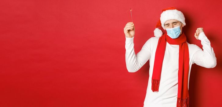 Concept of covid-19, christmas and holidays during pandemic. Happy man celebrating new year at party, wearing medical mask and santa hat, dancing with sparkler against red background.