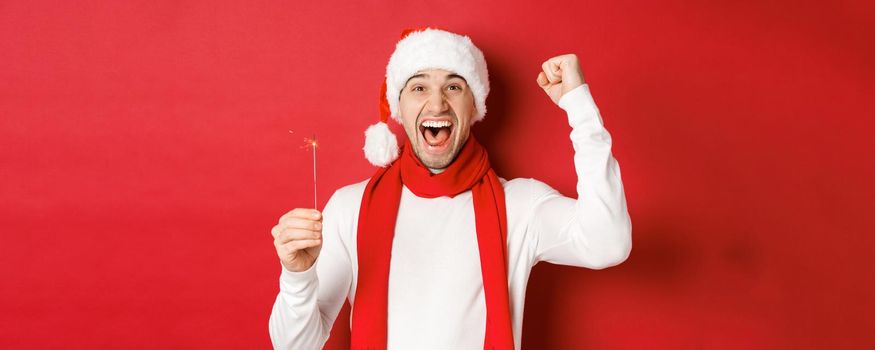 Concept of christmas, winter holidays and celebration. Portrait of excited handsome man, raising hand up and holding sparkler, wishing happy new year, standing over red background.