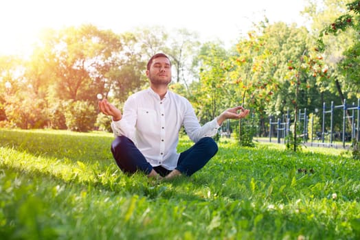 Young man meditates in lotus pose on green grass. Handsome man in casual wear practicing of yoga with closed eyes. Training and meditation outdoor at summer day. Healthy lifestyle and relaxation.