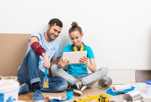 Happy couple watching something funny on tablet computer while sitting on floor. Painting tools and materials for home remodeling and interior redesign. Young family renovating their home after moving