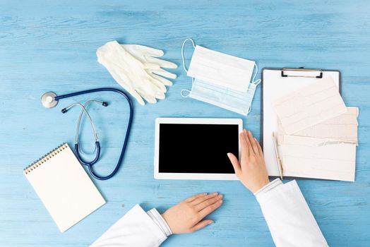 Top view of doctor hands with tablet computer. Cardiac examination in hospital. Therapist sitting at blue wooden desk with stethoscope, medical gloves and mask. Examination and consultation in clinic