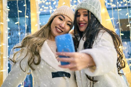 Two girlfriends asian girls in white coats are having fun hugging and selfie on mobile phone, new year, light bulbs