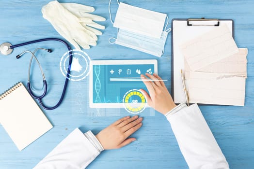 Top view of doctor hands with tablet computer. Therapist in white coat sitting at blue wooden desk. Examination and consultation in clinic. Medical application and online healthcare services.