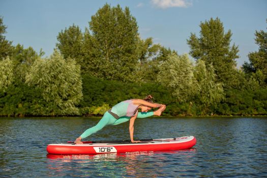 Young woman doing yoga on sup board with paddle. Yoga pose, side view - concept of harmony with the nature, free and healthy living, freelance, remote business.