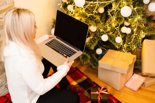 Pretty Young Woman in Trendy Winter Fashion Sitting on the Floor with Crossed Legs Holding Laptop Gadget in Front Christmas Tree Decoration, While Looking at the Camera.