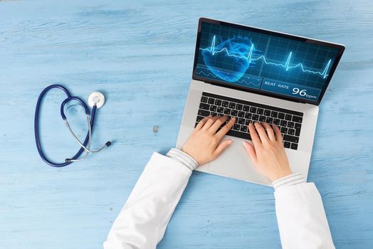 Top view of doctor hands typing at laptop computer. Therapist in white coat sitting at blue wooden desk. Online medical clinic communication with patient. Medical application and healthcare services.