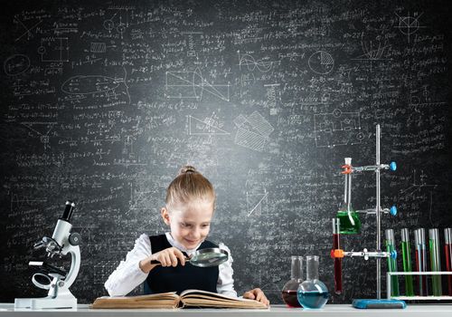 Little girl sitting at desk with magnifier and open book. Research and education in school. Happy schoolgirl in school wear carefully studying book. Clever girl reading book with magnifying glass.