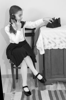 A beautiful little schoolgirl girl in a white blouse and black long skirt, with neatly braided pigtails on her head.She sits on an old Viennese chair and talks on an old telephone. Interior of the fifties of the last century.