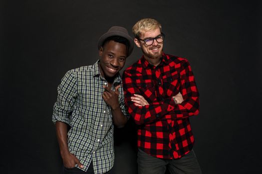 Studio shot of two stylish young men having fun. Handsome bearded hipster in a shirt in a cage standing next to his African-American friend in hat against a dark background. International friendship concept.