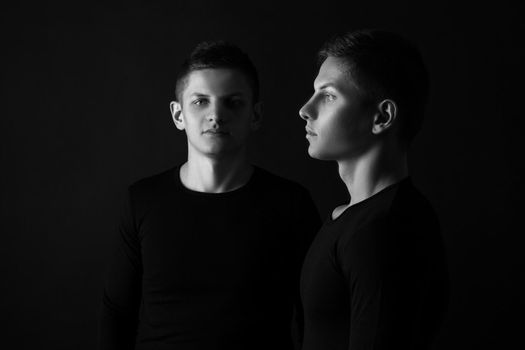 Two attractive positive young twin brothers standing dressed in black sweaters on black background. Black and white photography