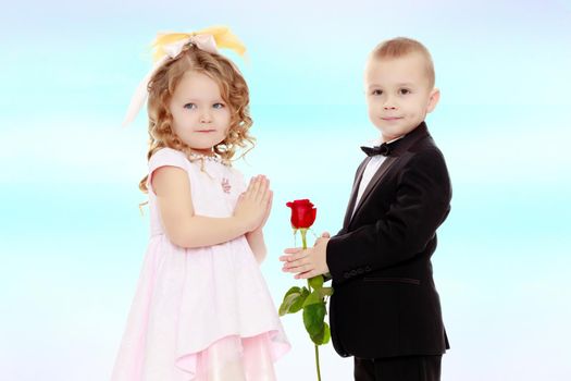 Little boy in black suit with bow tie gives a big red rose charming little girl.On the background of summer blue sky and fluffy clouds.