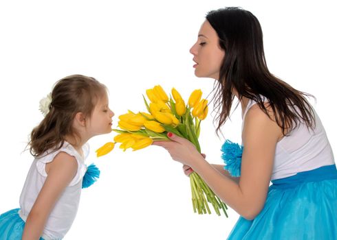 Beautiful young mother and little daughter in the same blue long skirts tutus , standing sideways to the camera ,admiring the bouquet of yellow tulips.Little girl enjoying the smell of flowers.Isolated on white background.