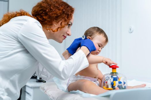 Red-haired female doctor examines little child for less than a year in a modern clinic. Pediatrician during examination baby in the hospital. Childrens healthcare. Diagnostics and treatment for kid.