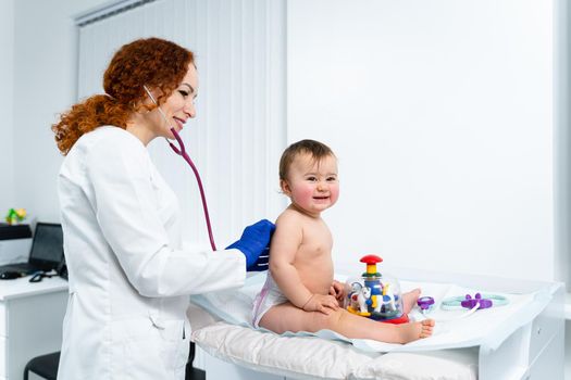 Red-haired female doctor examines little child for less than a year in a modern clinic. Pediatrician during examination baby in the hospital. Childrens healthcare. Diagnostics and treatment for kid.