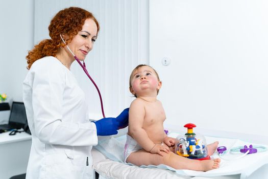 Red-haired female doctor examines little child for less than a year in a modern clinic. Pediatrician during examination baby in the hospital. Childrens healthcare. Diagnostics and treatment for kid.