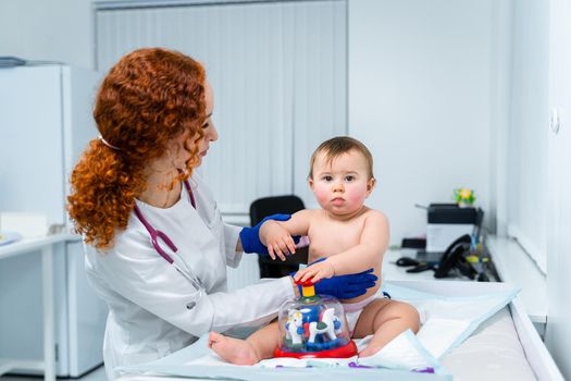 Red-haired female doctor examines little child for less than a year in a modern clinic. Pediatrician during examination baby in the hospital. Childrens healthcare. Diagnostics and treatment for kid.