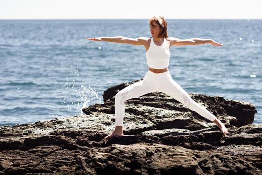 Young woman doing yoga in the beach. Female wearing white sport clothes in warrior 2 figure with defocused sea background.