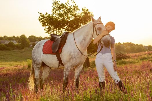 Taking care of animals, love and friendship concept. Jockey young girl petting and hugging white horse in evening sunset. Sun flare