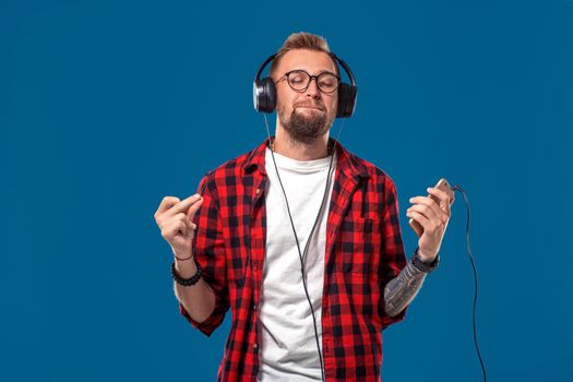 Happy young man listening to music with headphones. Handsome smiling guy in checkered shirt with closed eyes dancing with headphones. Isolated on blue background. Concept of people emotions