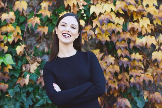 Beautiful young woman, model of fashion, wearing black dress smiling in urban background with autumn colors. Very straight hair styling