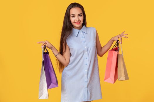 Image of a young brunette lady in white and blue summer striped dress posing with shopping bags and looking at camera over yellow background. Shopping. Sale