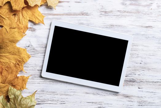 Tablet computer with blank screen lies on vintage wooden desk with bright foliage. Flat lay composition with autumn leaves on white wooden surface. Internet communication and digital technology