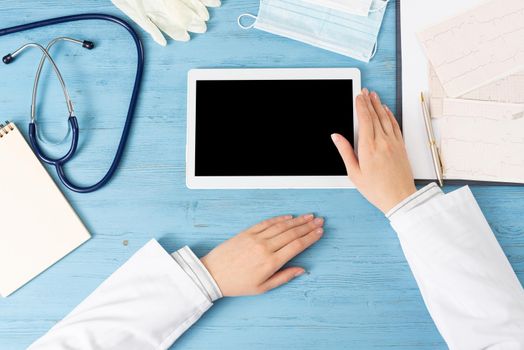 Top view of doctor hands with tablet computer. Cardiac examination in hospital. Therapist sitting at blue wooden desk with stethoscope, medical gloves and mask. Examination and consultation in clinic