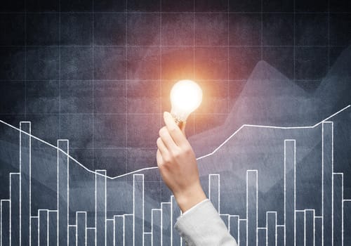 Businesswoman holding glowing light bulb on grunge background with financial diagram. Competent business data analysis. Idea generation and brainstorming. Successful solution concept with lamp.