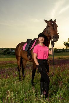 Beautiful smiling girl jockey stand next to her brown horse wearing special uniform on a sky and green field background on a sunset. Equitation sport competition and activity.