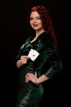 Young redheaded woman whith a curly hair in a black dress is posing with a playing cards in her hands and smiling, on black background Win casino poker slots