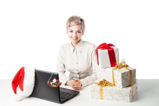 Pretty young lady sitting at desk with laptop present boxes and winter hat over white background