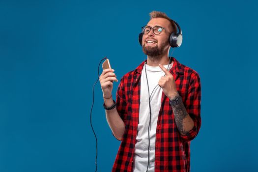 Dance Handsome young stylish man in headphones holding MP3 Player and dancing while standing against blue background. Emotional and people concept: young bearded man in checkered shirt. Hipster style. Copy space